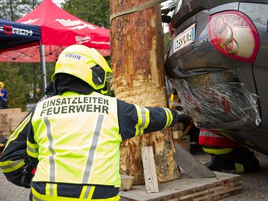Einsatzleiter Feuerwehr weist auf verunfalltes Auto, das gegen Baumstamm gepresst ist